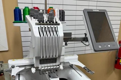 Brother six-needle embroidery machine with its touch screen on an arm in front of a ruled whiteboard