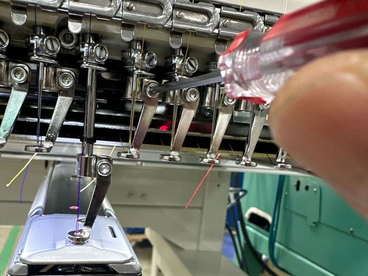 Hand with a red-handled driver adjusting a needle bar on a Tajima head