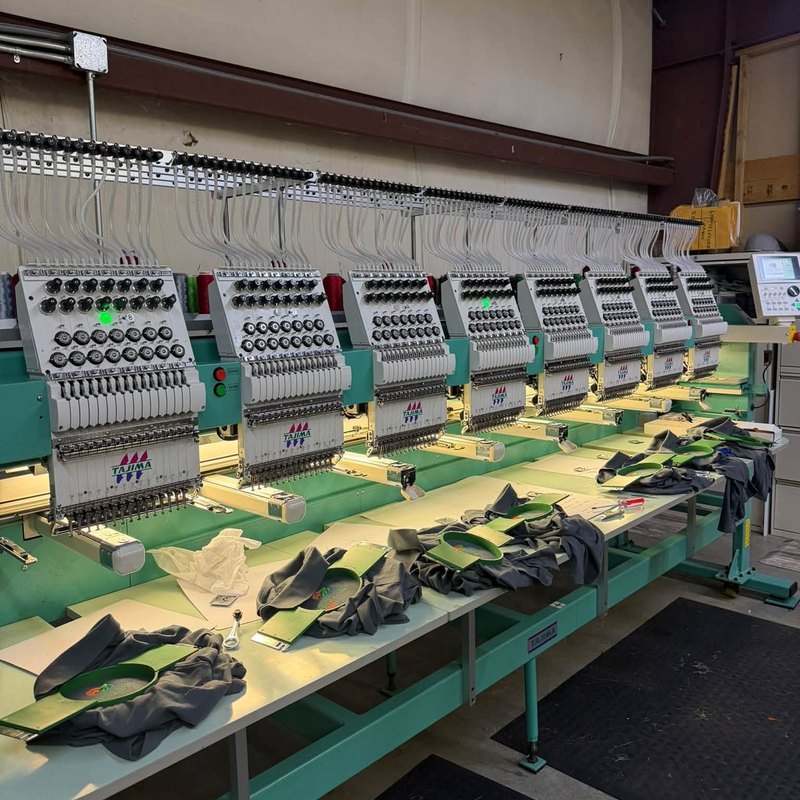 Eight-head Tajima embroidery machine with gray shirts hooped along the table and hand tools beside them