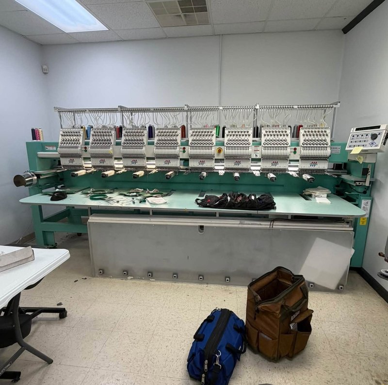 Eight-head Tajima embroidery machine with an open tool roll on its table and two tool bags on the floor